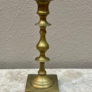 May include: A gold-toned brass candlestick with a square base and a tall, slender stem. The candlestick has a simple, elegant design.