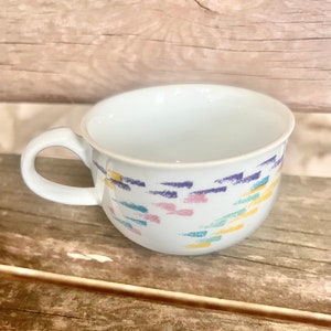 May include: White ceramic teacup with a colorful abstract pattern of yellow, blue, pink, and purple brushstrokes. The cup has a rounded shape and a handle.