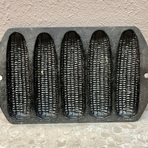 May include: A black cast iron corn-shaped cornbread pan with five corn cob molds. The pan has a rectangular shape with rounded edges and small holes on each end. The molds are detailed with textured kernels.