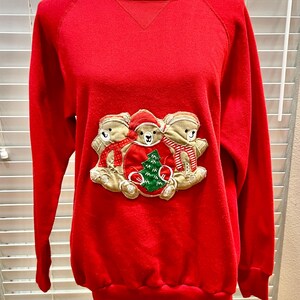 May include: A red sweatshirt with a crew neck and long sleeves. The front features an embroidered design of three teddy bears wearing scarves and hats, surrounding a Christmas tree. The bears are beige with red and green accents.