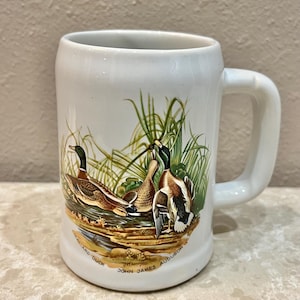 May include: White ceramic mug with a handle, featuring a detailed illustration of ducks in a marsh setting. The artwork includes green reeds, brown and orange ducks, and the text "MALLARD DUCK JOHN JAMES AUDUBON".