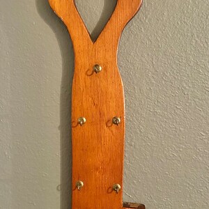 Puede incluir: Un colgador de llaves de madera con forma de llave con cinco ganchos de latón. El colgador de llaves está pintado de color marrón claro y tiene un acabado desgastado.