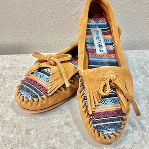 May include: A pair of tan suede moccasins featuring a multi-colored woven fabric insert with geometric patterns, fringe detailing, and a decorative bow on the vamp. The interior insole displays the Minnetonka brand label on a striped fabric.