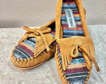 May include: A pair of tan suede moccasins featuring a multi-colored woven fabric insert with geometric patterns, fringe detailing, and a decorative bow on the vamp. The interior insole displays the Minnetonka brand label on a striped fabric.