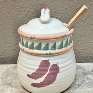 May include: A white ceramic sugar bowl with a lid and a small wooden spoon. The bowl features a pink and green geometric design around the middle and two purple chili pepper illustrations. The lid has a small handle.