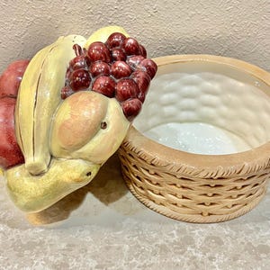 May include: A ceramic fruit basket with a detailed design. The basket is tan with a woven pattern. A cluster of grapes, bananas, peaches, and apples are sculpted on the side. The fruit is painted in realistic colors.