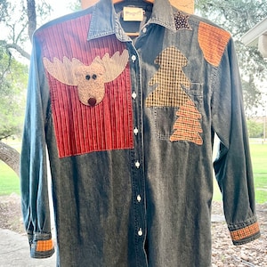 May include: A blue denim shirt with patchwork details. The patches feature a moose, two Christmas trees, and a red and white striped fabric. The shirt has long sleeves and a button-up front.
