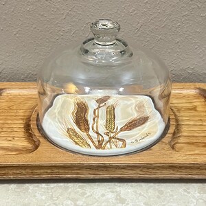 May include: A wooden serving tray with a clear glass dome and a decorative plate featuring wheat stalks. The tray has two recessed areas and a light brown finish. The plate has a white background with wheat stalks in shades of gold and brown.