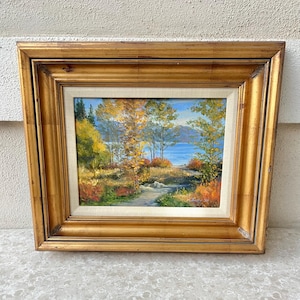May include: A framed oil painting depicting a landscape scene with a lake, trees, and a path leading to the water. The painting is in a gold frame and has a rustic feel.