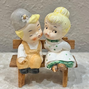 May include: A vintage ceramic figurine set depicting a boy and girl seated on a wooden bench. The boy wears a gray hat and overalls, while the girl is in a white dress with green trim. The figurines are painted with soft colors and have a nostalgic charm.
