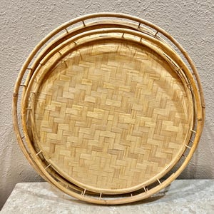 May include: A set of three round woven wicker trays. The trays are nested inside each other, with the largest tray on the bottom and the smallest tray on top. The trays are made of natural woven wicker and have a light brown color.