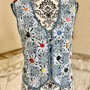May include: A light blue crochet vest with a daisy flower pattern. The vest has a V-neck and is adorned with colorful buttons in red, yellow, orange, and black. The vest is displayed on a mannequin.
