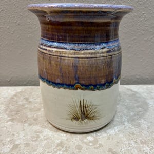 May include: A handcrafted ceramic vase with a wide, flared rim. The upper portion is a rich brown with blue and tan accents, transitioning to a white base. A detailed illustration of a plant is on the lower portion.