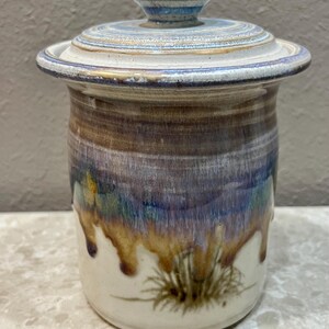 May include: A handcrafted ceramic jar with a lid. The jar features a cylindrical shape with a white base and a gradient of brown, blue, and gold hues. The lid is adorned with concentric blue and white stripes. The jar has a rustic, artisanal aesthetic.