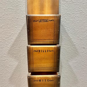 May include: A vertical wooden wall organizer featuring three stacked storage compartments. Each compartment is labeled with black text reading LETTERS, BILLS, and MISC. The wood has a medium brown finish with darker edges and a rounded top.