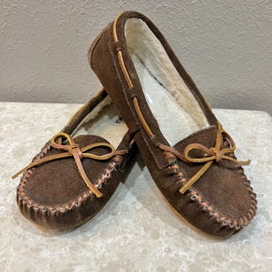 May include: A pair of brown suede moccasin slippers. The slippers have a soft, light-colored lining and are adorned with leather ties and decorative stitching around the edges. The design is classic and comfortable.