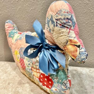 May include: A stuffed fabric bird figure crafted from a patchwork quilt featuring various multicolored floral and geometric patterns. The bird has a prominent pink fabric head and is adorned with a decorative blue satin ribbon bow around its neck.
