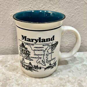 May include: A cream-colored ceramic mug with a dark teal interior and a black rim. The exterior features a black line-art map of Maryland, including the text Maryland, Cumberland, Elkton, Frederick, Baltimore, Annapolis, Leonardtown, Easton, and Ocean City.