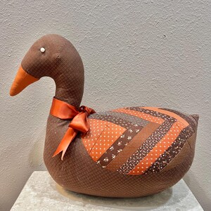 May include: A brown fabric duck with an orange beak and a decorative orange ribbon tied around its neck. The duck's body features a patchwork design with various brown and orange patterned fabrics. A decorative accent for a home.