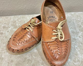 May include: A pair of tan leather huarache-style shoes featuring a woven upper design, light-colored laces, and white stitching. The interior of the shoe is stamped with the number 17 and the brand name Torry accompanied by a turtle graphic.