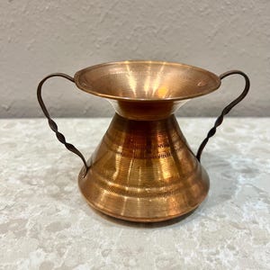 May include: A small, copper-colored vase with a wide, shallow top and a flared base. The vase has two twisted handles and horizontal etched lines around the body. The vase is likely made of copper or a copper alloy.