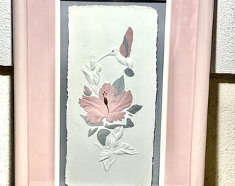 May include: A framed pink and white embossed paper print of a hummingbird hovering near a pink flower with green leaves.