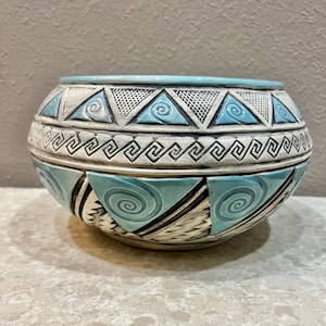May include: A ceramic bowl with a rounded shape, featuring a light blue rim and geometric patterns. The bowl's design includes triangles, spirals, and a repeating Greek key pattern in shades of white, black, and turquoise. The bowl sits on a light-colored surface.