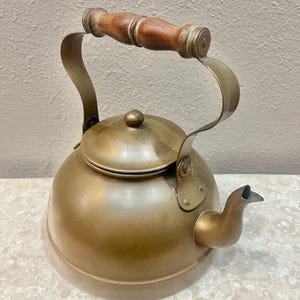 May include: A vintage brass kettle with a wooden handle. The kettle has a rounded body, a curved spout, and a lid with a small knob. The handle is made of wood and is attached to the kettle with brass fittings. A classic kitchenware item.