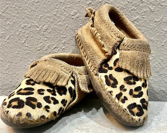 May include: A pair of tan suede baby moccasins with a leopard print design. The shoes have a fringed trim and a soft sole. The moccasins are designed for infants and toddlers.