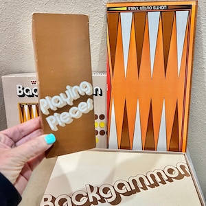 May include: A brown box labeled Playing Pieces held in front of a backgammon game set. The set includes a board with orange and white triangular points, a box with the text Backgammon, and a box showing a game board with yellow and brown checkers.