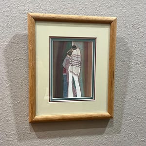 May include: Framed artwork featuring a stylized depiction of two figures embracing. The artwork uses muted tones of brown, red, and blue, with a focus on the figures' clothing and posture. The frame is made of light-colored wood, adding a natural touch.