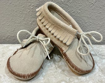 May include: A pair of beige suede moccasins with fringe detailing and brown stitching. The shoes have white laces and are designed for infants or toddlers. The soft, neutral colors and classic design make them a versatile choice.