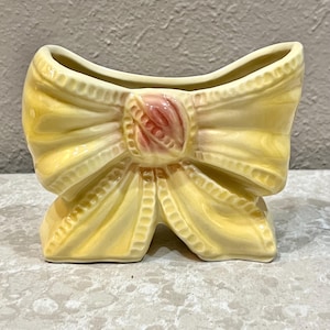 May include: A ceramic vase shaped like a bow, in a soft yellow color. The bow has detailed ridges and a pink accent in the center. The vase is designed with a curved opening at the top.