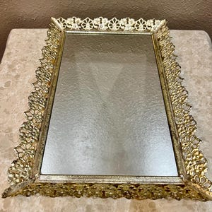 May include: A rectangular gold-toned vanity tray with a mirrored surface. The tray features an ornate, openwork design around the edges. The decorative border has a repeating floral pattern. The tray is likely made of metal.