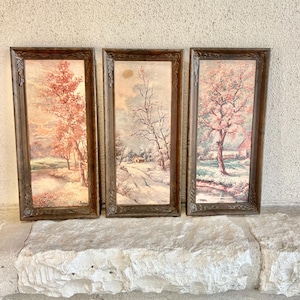 May include: Set of three framed landscape paintings. Each painting depicts a different season with trees, a house, and a stream. The frames are dark brown with ornate detailing. The paintings feature soft colors of pink, white, and brown.
