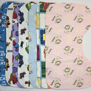 May include: Seven different fabric bibs with various children's book character prints. The bibs are all white with different colored prints. The prints include The Cat in the Hat, Brown Bear, Brown Bear, What Do You See?, Where the Wild Things Are, and more.