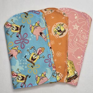May include: Three fabric items featuring SpongeBob SquarePants and related characters. The first item is light blue with SpongeBob and Patrick Star, the second is orange with SpongeBob, and the third is pink with SpongeBob and other characters. The items have rounded edges.
