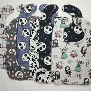 May include: Six baby bibs with different patterns, featuring characters and motifs from "The Nightmare Before Christmas." The colour scheme includes black, white, grey, and hints of purple and green. The bibs are designed for babies and young children.