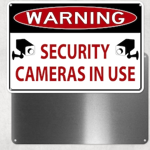 May include: A rectangular metal sign with the words "WARNING" in a red oval at the top. Below, the sign reads "SECURITY CAMERAS IN USE" in red text, with black security camera icons on either side.