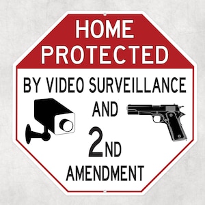 May include: A red and white octagonal sign with the words "HOME PROTECTED" at the top. Below, it reads "BY VIDEO SURVEILLANCE AND 2ND AMENDMENT." The sign features a security camera and a handgun.