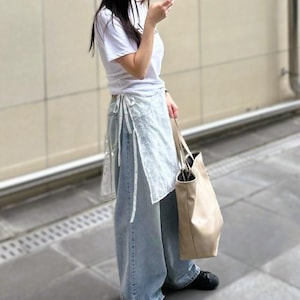 Peut inclure: Une personne porte un t-shirt blanc, un jean bleu clair et des chaussures noires. Elle tient un sac fourre-tout beige et un smartphone. La tenue est décontractée et adaptée au quotidien.