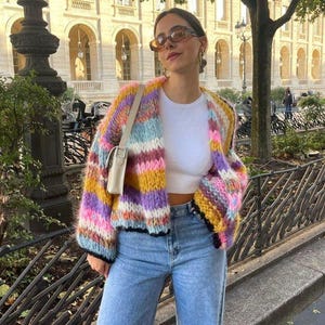 Könnte beinhalten: Ein farbenfroher, gestreifter Strickcardigan in Gelb-, Rosa-, Lila- und Blautönen. Der Cardigan wird über einem weißen Oberteil und mit hellblauen Jeans getragen. Eine kleine, cremefarbene Handtasche wird über der Schulter getragen.