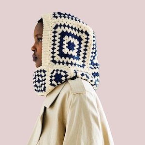 May include: A white and blue crocheted hooded scarf with a geometric pattern. The scarf is worn over a beige trench coat.