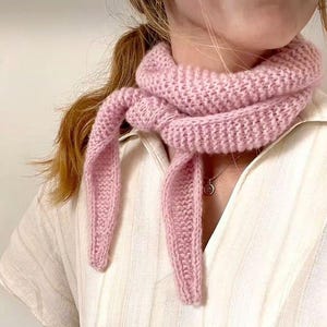 May include: A pale pink knitted scarf, looped around the neck and tied. The scarf is made of a thick, textured yarn. The background is a white shirt with vertical stripes. A silver necklace is visible.