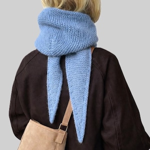 Op de afbeelding: Een lichtblauwe gebreide sjaal met lange, puntige uiteinden. De sjaal is om de nek gewikkeld en over een bruine jas gedragen. Een beige schoudertas is zichtbaar. De sjaal is een zachte, getextureerde brei.