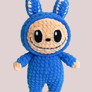Può includere: Un giocattolo di peluche blu fatto a mano a forma di creatura con un viso sorridente, grandi occhi neri e piccoli denti. Il giocattolo ha un completo blu con cappuccio con orecchie abbinate e accenti beige.