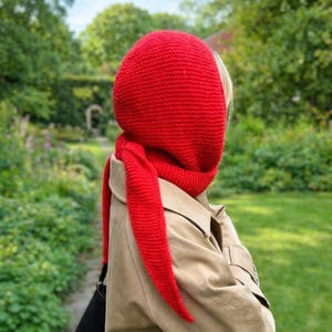 Könnte beinhalten: Ein leuchtend rotes Strick-Set aus Kapuze und Schal. Die Kapuze bedeckt Kopf und Hals, der Schal verläuft über die Vorderseite. Die Person trägt einen beigefarbenen Trenchcoat und eine schwarze Tasche. Der Hintergrund ist ein grüner Garten.