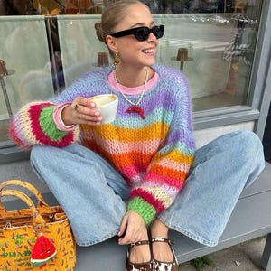 Op de afbeelding: Een persoon draagt een regenboog gestreepte trui, lichtblauwe jeans en schoenen met koeienprint. Een gele handtas met een watermeloen bedel staat in de buurt. De persoon houdt een kop koffie vast en draagt een zonnebril.