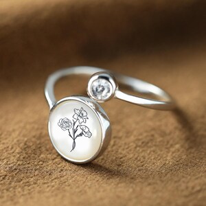 May include: A silver-toned ring with a floral design. The ring features a round, white face with a black floral illustration. A small, clear gemstone is set on the band. The ring is set against a brown background.