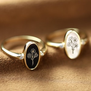 以下が含まれることがあります： 楕円形のセッティングが施された金のリングが2つ。1つのリングは、黒いセッティングに金のフローラルデザインが施されています。もう1つのリングは、白いセッティングに金のフローラルデザインが施されています。